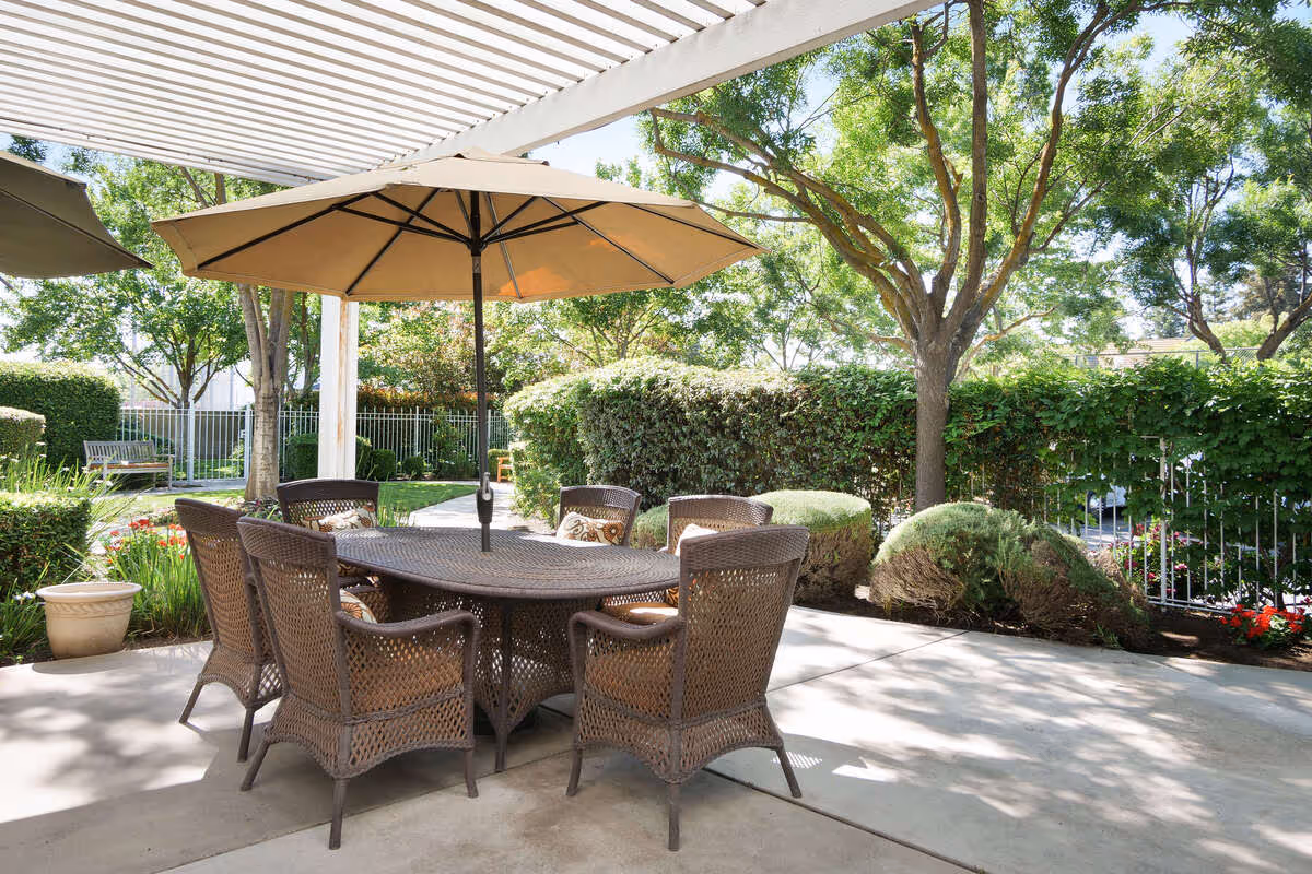 Outdoor patio area with a round wicker table and six matching wicker chairs, shaded by a large beige umbrella. The patio is surrounded by green trees, bushes, and a white fence, with sunlight filtering through the leaves.