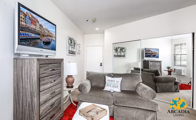 A cozy living room with a gray sofa featuring a decorative pillow that says 'Home is where we are.' A wooden dresser with a flat-screen TV mounted above it is on the left side. A small round table with a lamp is next to the sofa. The room has a large mirrored closet door reflecting the furniture and a window with blinds. A red rug is partially visible on the floor. The Arcadia Living logo is in the bottom right corner.