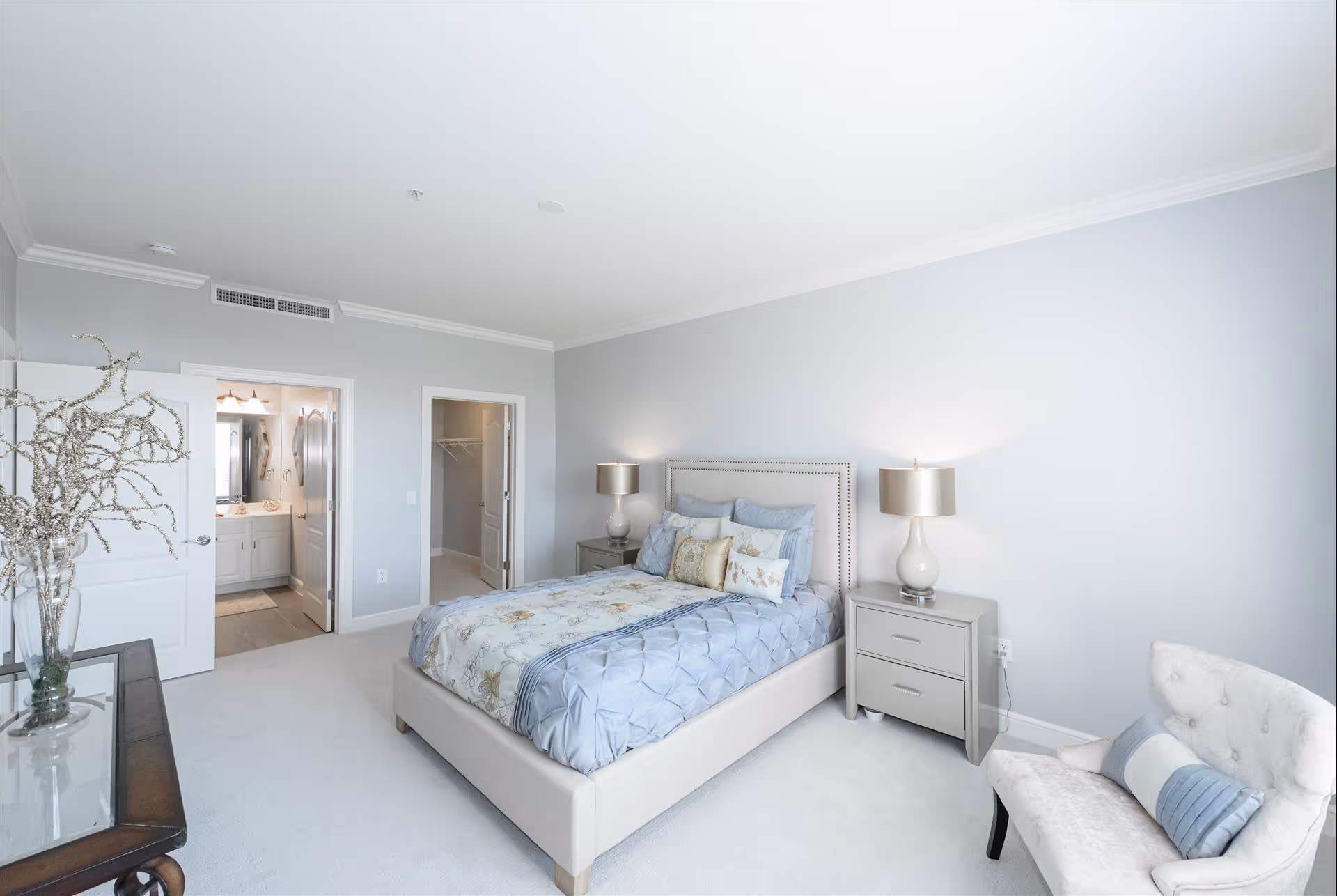Bright, neutral master bedroom with an upholstered bed, matching nightstands and lamps, an armchair, and open doors to a bathroom and walk-in closet.