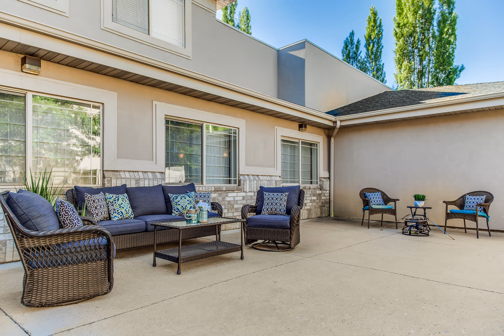 Outdoor patio with wicker sofas and chairs topped with blue cushions and throw pillows in front of a beige building.