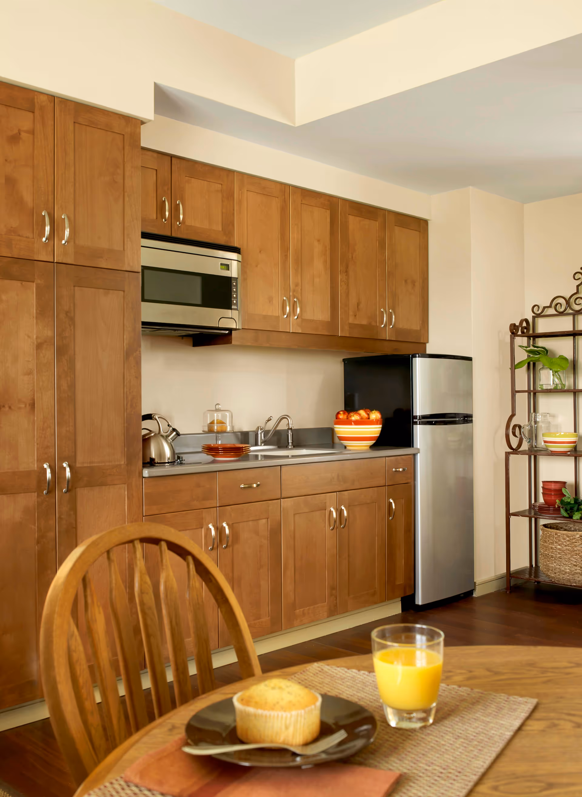 A kitchen area with wooden cabinets, a stainless steel microwave, a small refrigerator, and a countertop with a sink. On the counter, there is a bowl of apples and a kettle. In the foreground, there is a wooden dining table with a muffin on a plate and a glass of orange juice.