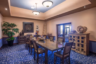 A well-decorated dining room with a long wooden table surrounded by eight chairs. The room features a patterned blue carpet, beige walls with a chair rail, and a tray ceiling with two ceiling lights. There is a large plant in the corner, decorative items on a sideboard and a cabinet, and a glass door leading to another room.