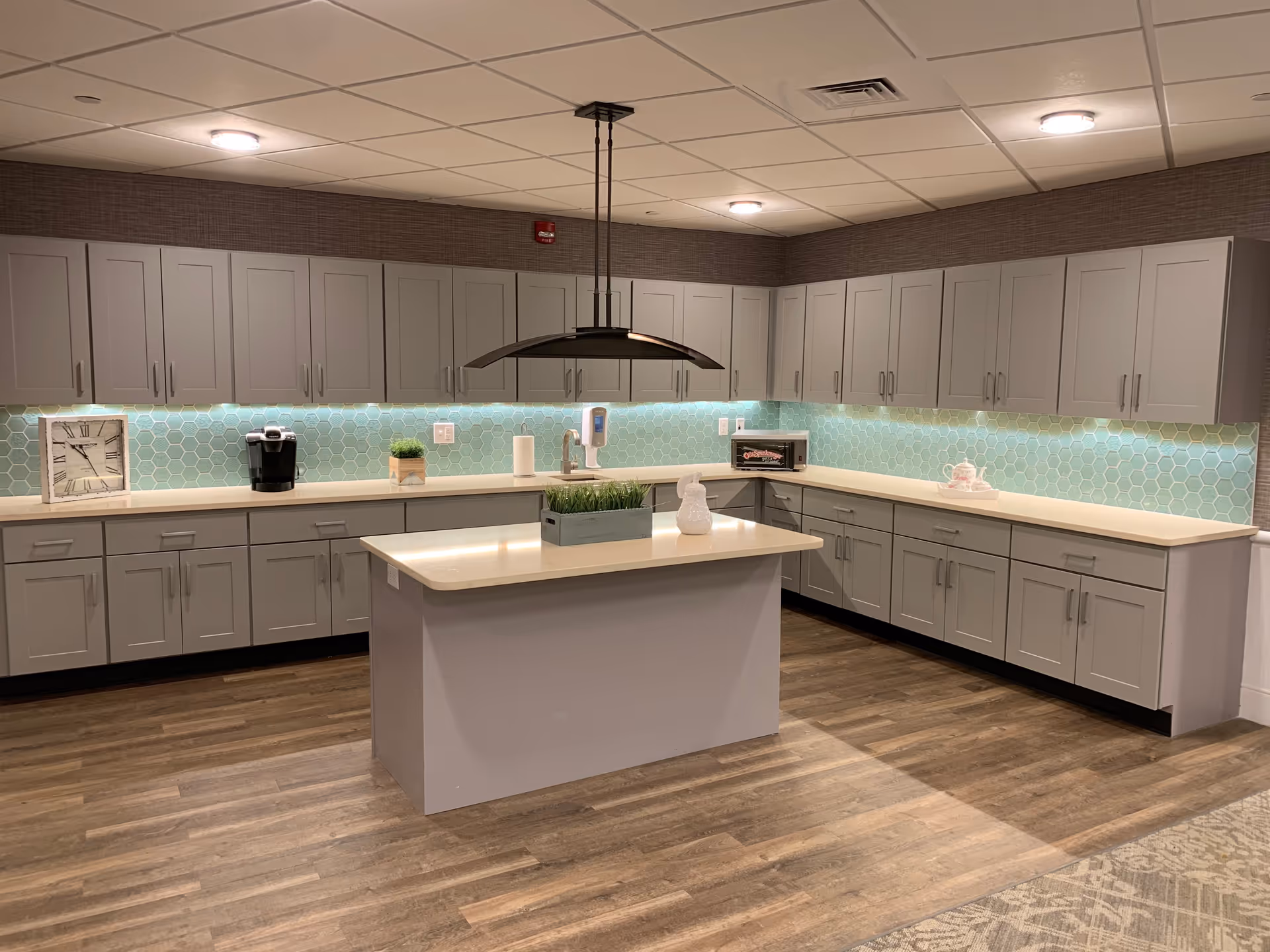 A modern kitchen area with light gray cabinets and drawers lining the walls and a central island with a light countertop. The backsplash features light blue hexagonal tiles with under-cabinet lighting. On the countertop, there is a clock, a coffee maker, a small plant, a paper towel roll, a soap dispenser, a toaster oven, and a decorative white ceramic duck. The floor is wood-patterned, and the ceiling has recessed lighting and a hanging light fixture above the island.