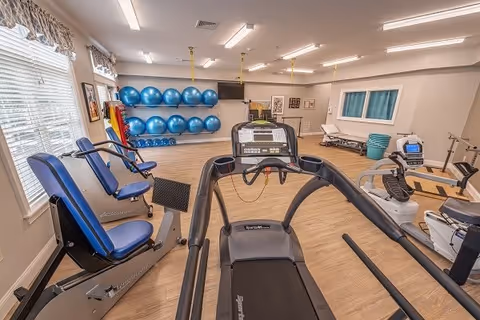 A well-lit fitness room in a senior living facility featuring exercise equipment including a treadmill, stationary bikes, and blue padded chairs. The room has wooden flooring, large windows with blinds, and a wall-mounted rack holding multiple blue exercise balls. There are framed pictures on the walls and a treatment table in the back corner.