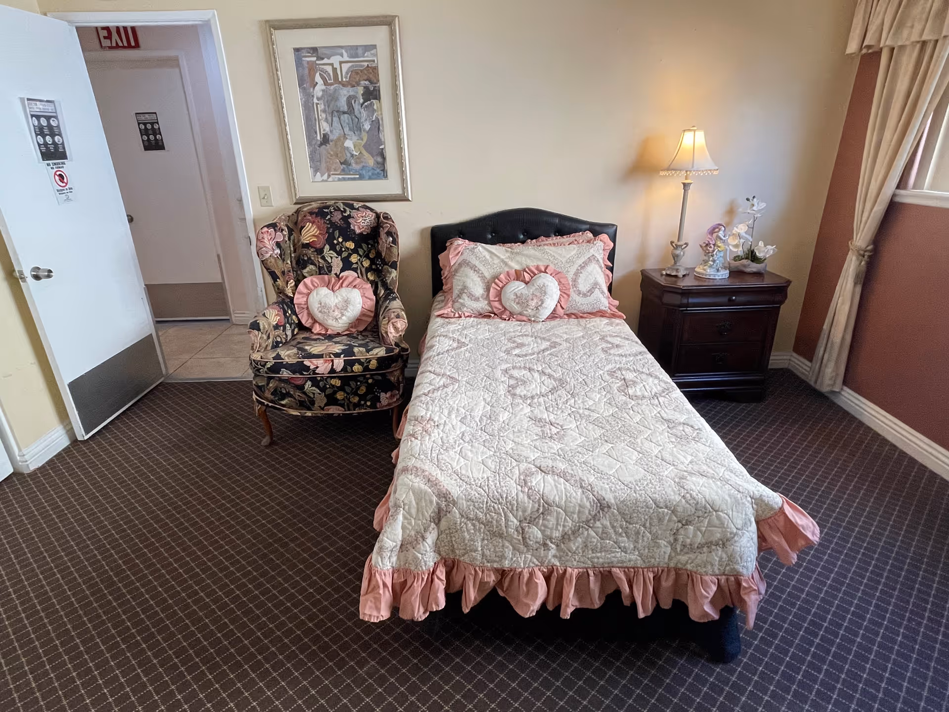 A cozy bedroom with a single bed covered in a quilted bedspread with pink ruffles and heart-shaped pillows. Next to the bed is a dark wooden nightstand with a lamp and decorative items. A floral upholstered armchair with a matching heart-shaped pillow is positioned beside the bed. The room has carpeted flooring, a window with curtains, and an open door leading to another room.