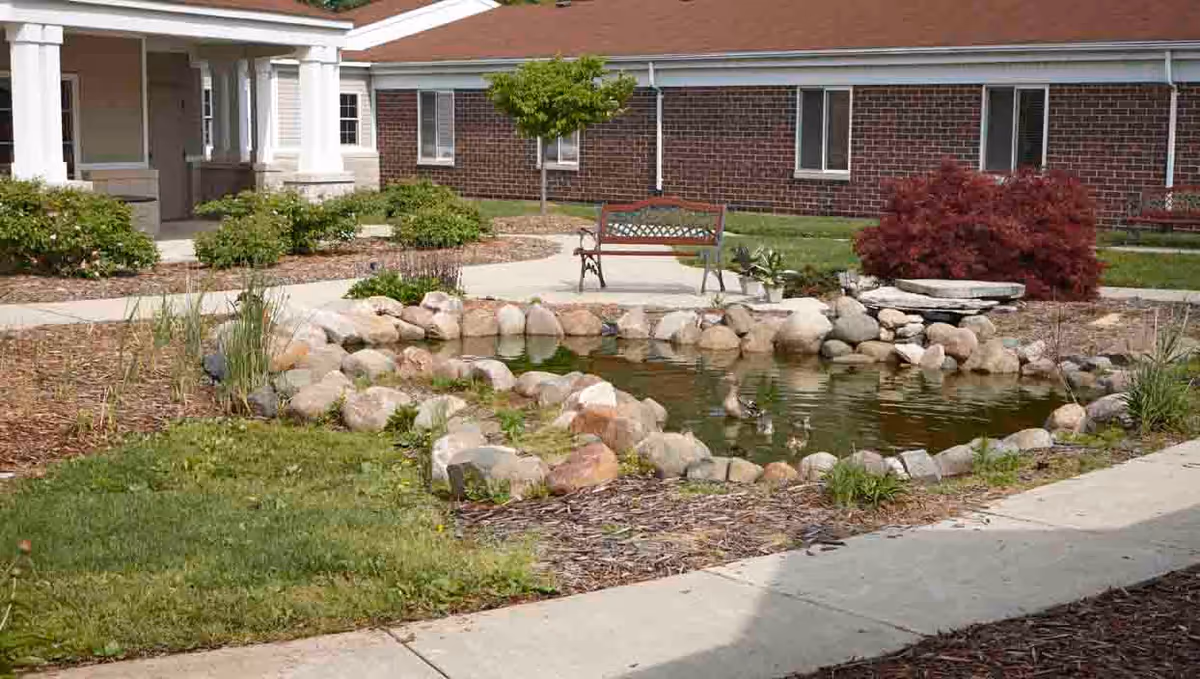 Outdoor courtyard area at The Lodge at Taylor featuring a small pond surrounded by rocks, a wooden bench, green grass, shrubs, and a brick building with white trim in the background.