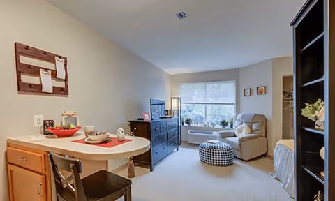 A cozy living area in a senior living facility featuring a small dining table with two chairs, a dresser with a television on top, a comfortable armchair with a footstool, and a large window letting in natural light. The room has neutral-colored walls and carpeted flooring, with decorative items on the walls and shelves.