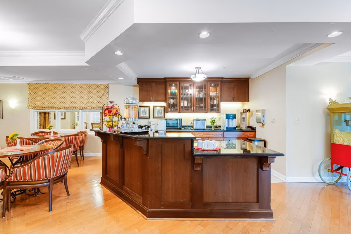 Interior view of a senior living facility kitchen and dining area featuring a large wooden island with a black countertop, a beverage dispenser, and a tray with cups. Behind the island are wooden cabinets with glass doors, a coffee machine, and other kitchen appliances. To the left, there are dining tables with striped upholstered chairs and a window with a patterned valance. On the right side, there is a popcorn machine with a red base and yellow top on wheels. The floor is wooden, and the room is well-lit with ceiling lights.