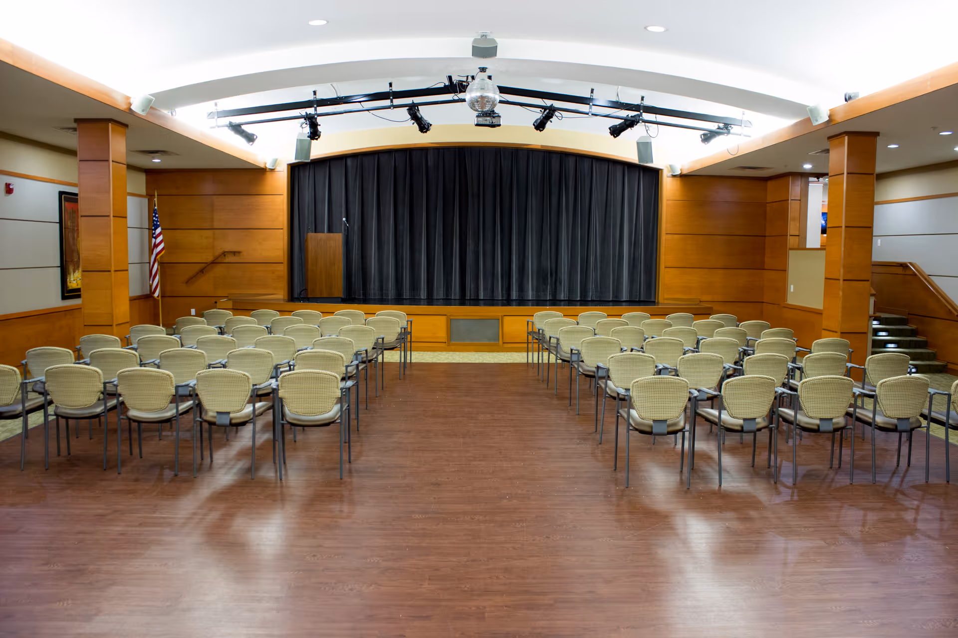 An empty auditorium or meeting room with rows of beige chairs facing a stage with a closed black curtain. The room has wooden paneling on the walls, an American flag on the left side, and overhead stage lighting and a disco ball above the stage.