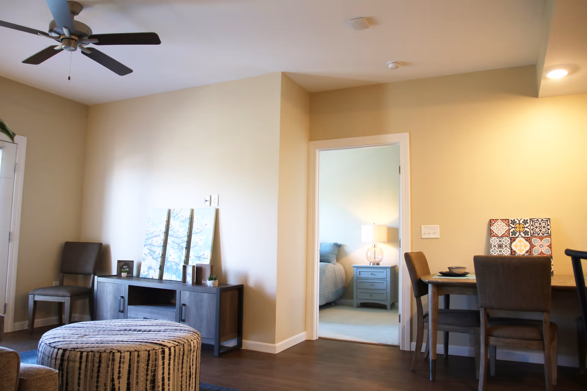 Open living room with an ottoman, media console, dining table and chairs, and a view into a bedroom through a doorway.
