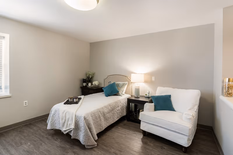 A bright, simply furnished bedroom with a single bed, bedside table and lamp, and a white armchair with teal pillows.
