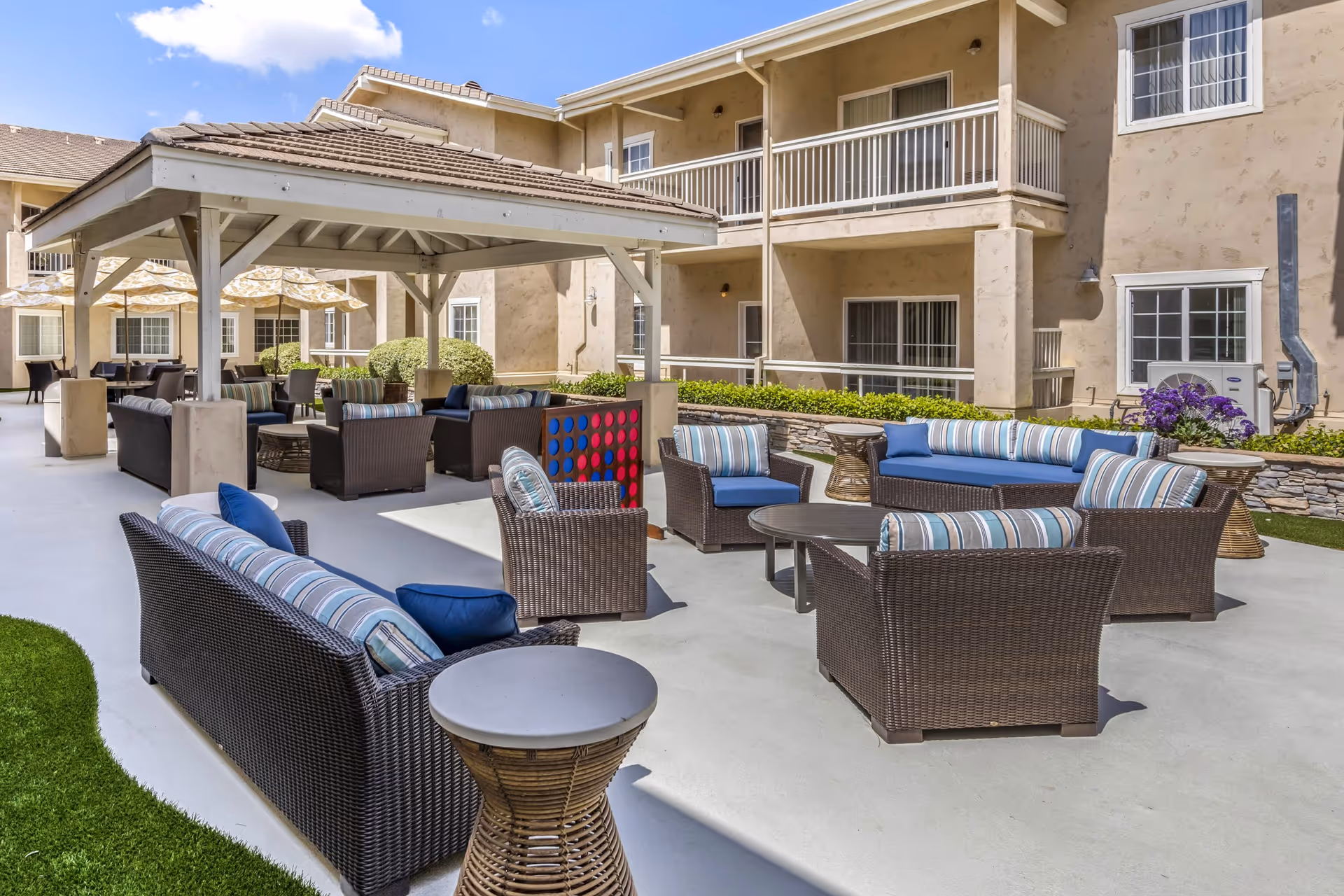 Outdoor patio area at Meadowbrook At Agoura Hills featuring multiple wicker sofas and chairs with blue and striped cushions arranged around small tables. A covered gazebo with additional seating is visible, along with a large Connect Four game. The building exterior with balconies and windows surrounds the patio under a partly cloudy sky.