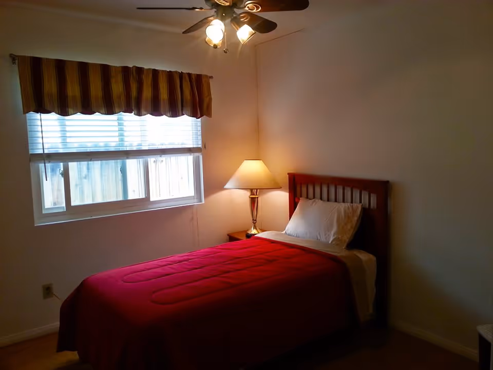 Small bedroom with a single bed covered by a red bedspread, a bedside lamp, window with blinds and a striped valance, and a ceiling fan.