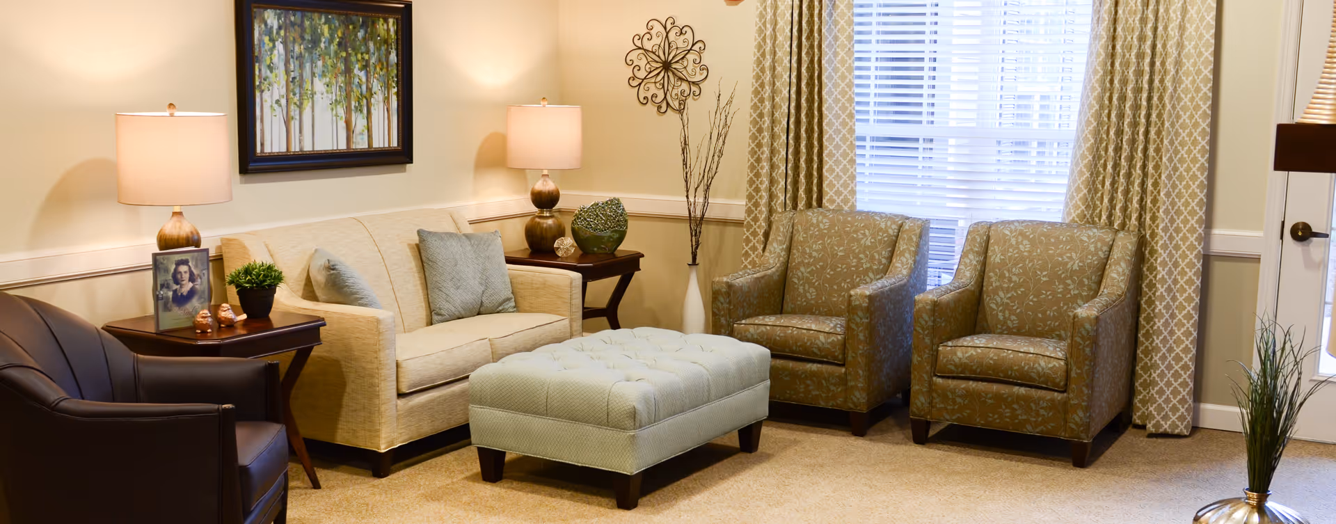 A cozy living room with a beige sofa, two patterned armchairs, a dark leather chair, and a large tufted ottoman in the center. The room is softly lit by two table lamps on side tables, with decorative items including a framed photo, a small plant, and a green vase. A large window with patterned curtains lets in natural light, and a framed painting of trees hangs on the wall above the sofa.