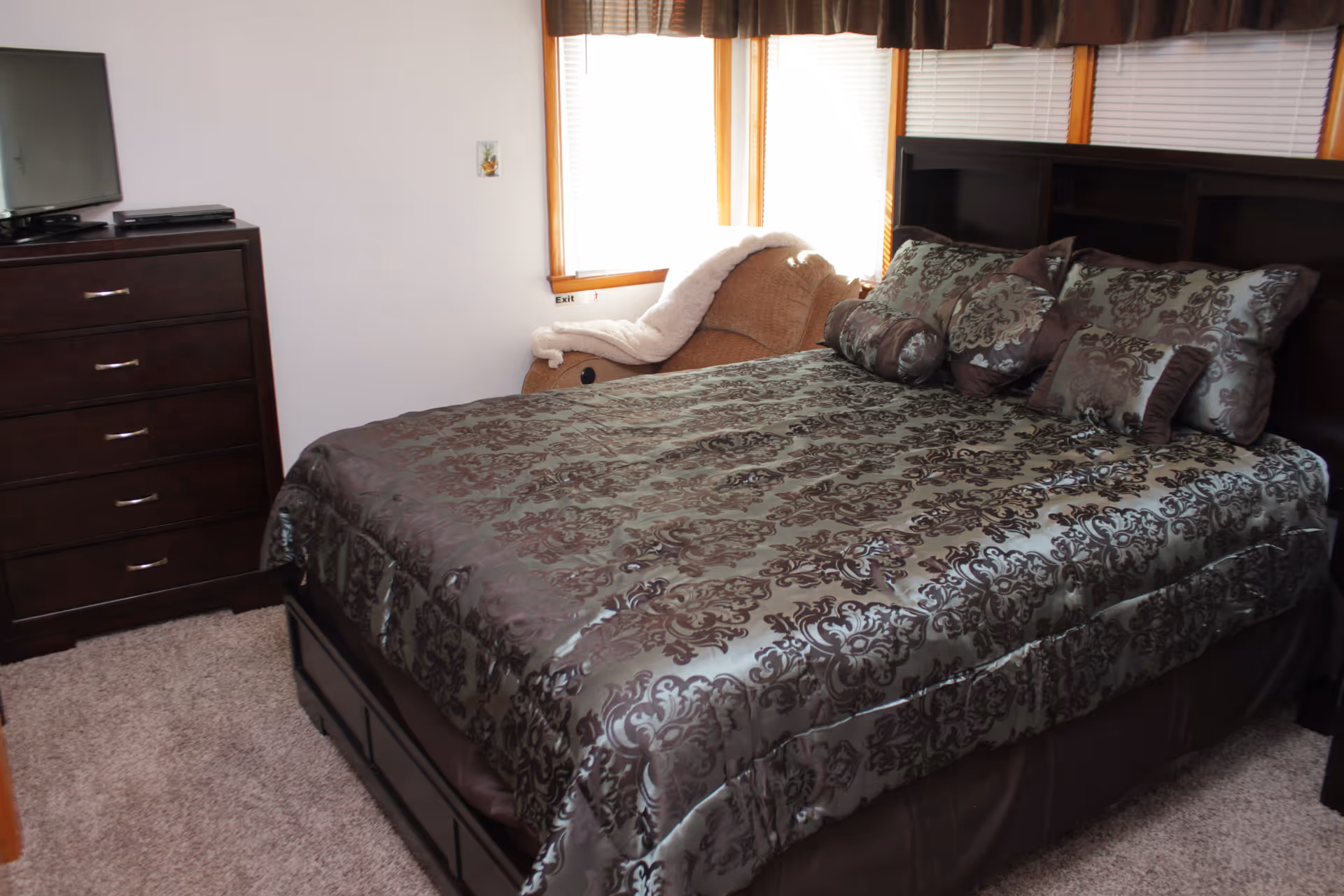 A bedroom with a large bed covered in a patterned brown and gray comforter with matching pillows. There is a dark wooden dresser with a flat-screen TV on top to the left and a beige armchair with a white blanket draped over it near the window. The room has carpeted flooring and white walls.