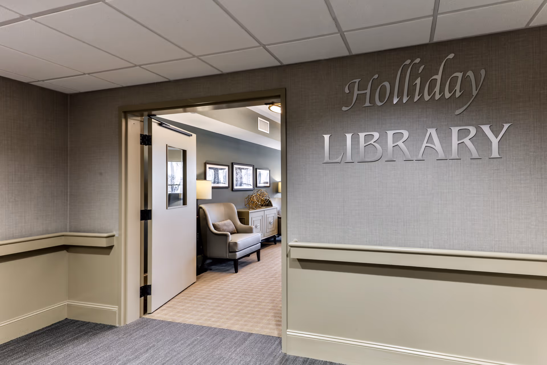 View of an open door leading into the Holliday Library room in a senior living facility. The hallway walls are beige with a chair rail, and the library room has a comfortable armchair, a sideboard with decorative items, and framed pictures on the wall.