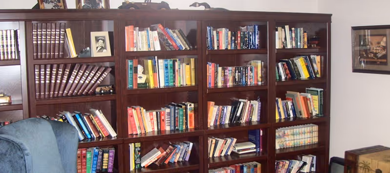 A cozy room featuring a large wooden bookshelf filled with various books and a framed black and white photo on one shelf. There is a blue upholstered chair partially visible in the foreground and a framed picture hanging on the wall to the right.