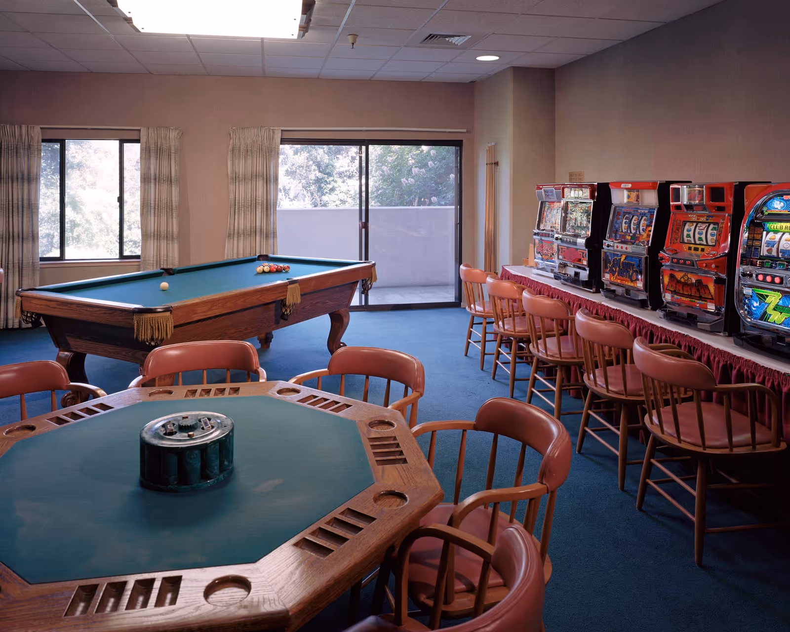 A recreational room with a pool table, an octagonal card table with chairs, and a row of slot machines along one wall. The room has large windows with curtains and a sliding glass door leading to an outdoor area.