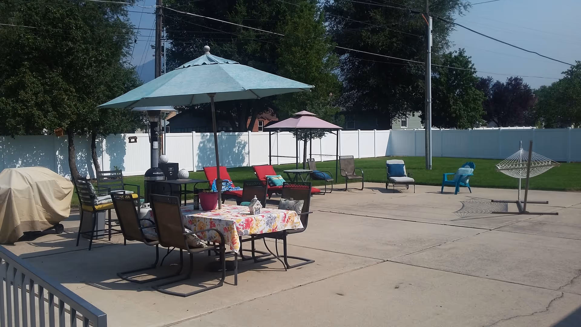 Outdoor patio area with multiple seating options including chairs, tables with umbrellas, lounge chairs, and a hammock stand on a concrete surface next to a grassy lawn enclosed by a white fence.