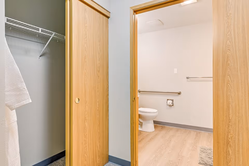 View of a bathroom with a toilet, grab bars, and a toilet paper holder, seen through an open wooden sliding door. To the left, there is a closet with a white robe hanging and a wire shelf inside.