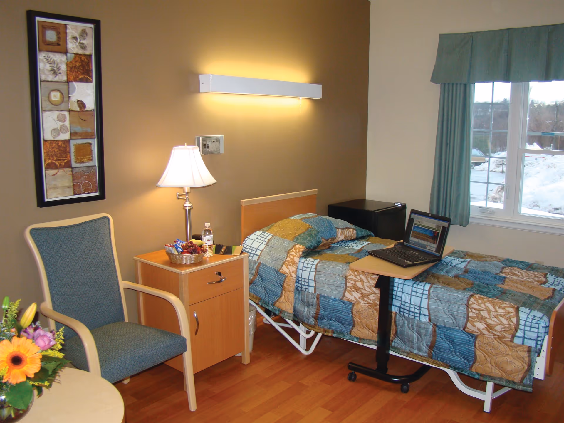 A small bedroom with a single bed covered in a patchwork quilt. Next to the bed is a wooden nightstand with a lamp, a basket of snacks, and a water bottle. A green upholstered chair is beside the nightstand. A laptop is placed on a rolling overbed table on the bed. A window with green curtains shows a snowy outdoor scene.