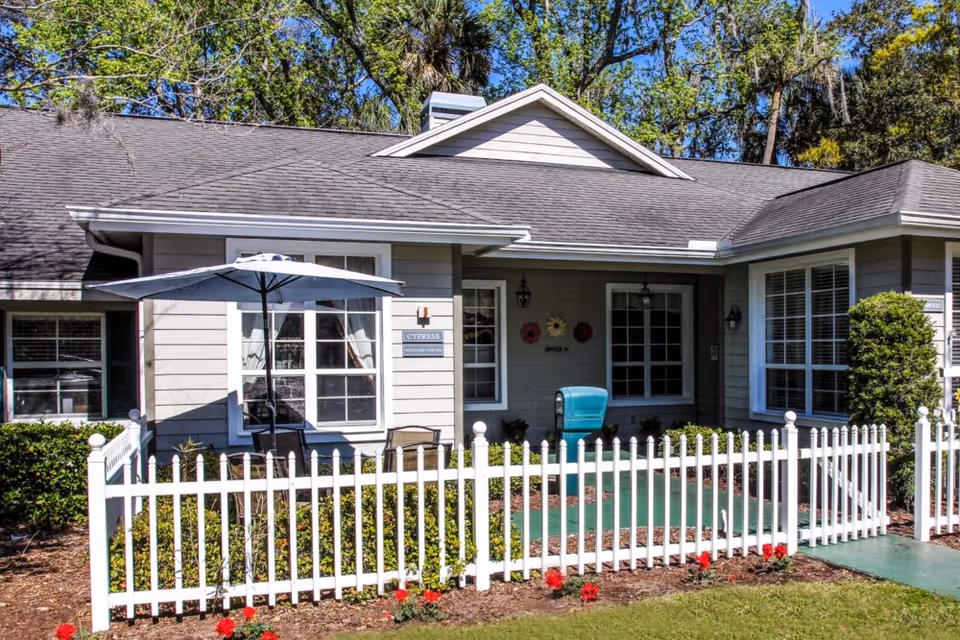 Exterior view of a single-story residential building with gray siding and a dark shingled roof. The front yard is enclosed by a white picket fence with a small garden bed featuring red flowers. There is a patio area with a table, chairs, and a large umbrella. Trees and greenery are visible in the background under a clear blue sky.