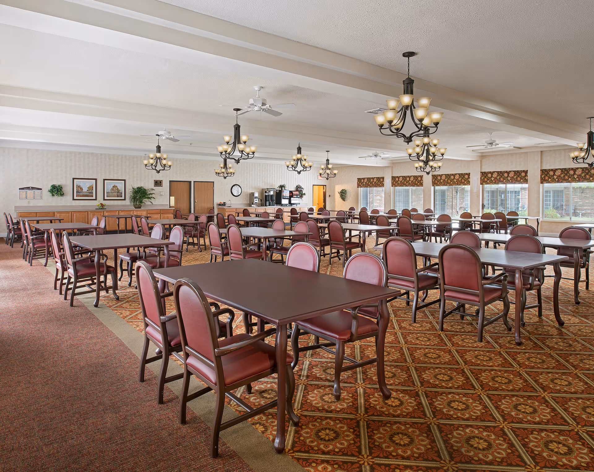 A spacious dining room with multiple dark wood tables and red cushioned chairs arranged neatly. The room features patterned carpet flooring, several chandeliers hanging from the ceiling, large windows with floral valances allowing natural light, and a beverage station against the far wall.