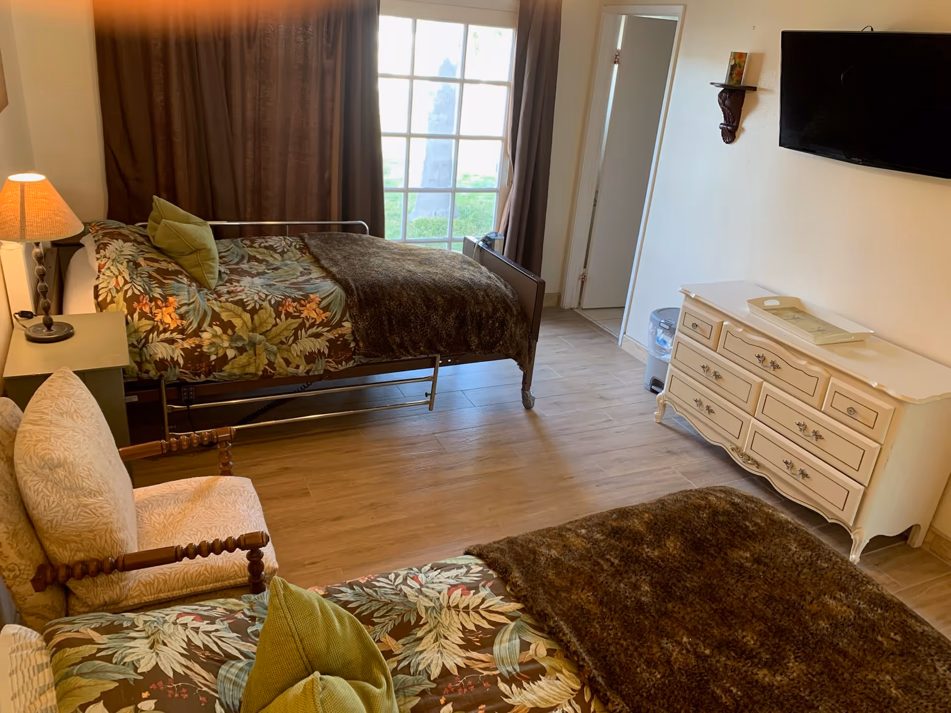A bedroom with two beds covered in floral patterned bedspreads and brown blankets. There is a cushioned wooden chair with a pillow, a bedside table with a lamp, a white dresser with a tray on top, a wall-mounted TV, and a large window with brown curtains letting in natural light. The floor is wooden, and there is a door leading to another room.