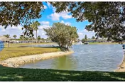 A peaceful outdoor scene featuring a small lake or pond surrounded by grassy banks and trees under a partly cloudy blue sky.