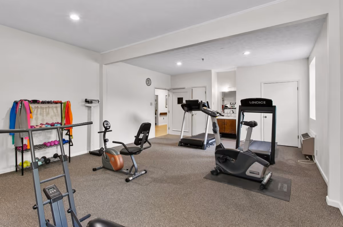 A fitness room in a senior living facility with exercise equipment including a treadmill, stationary bike, recumbent bike, and a rack of colorful dumbbells and resistance bands against a white wall. The room has beige carpet, white walls, and recessed ceiling lights.