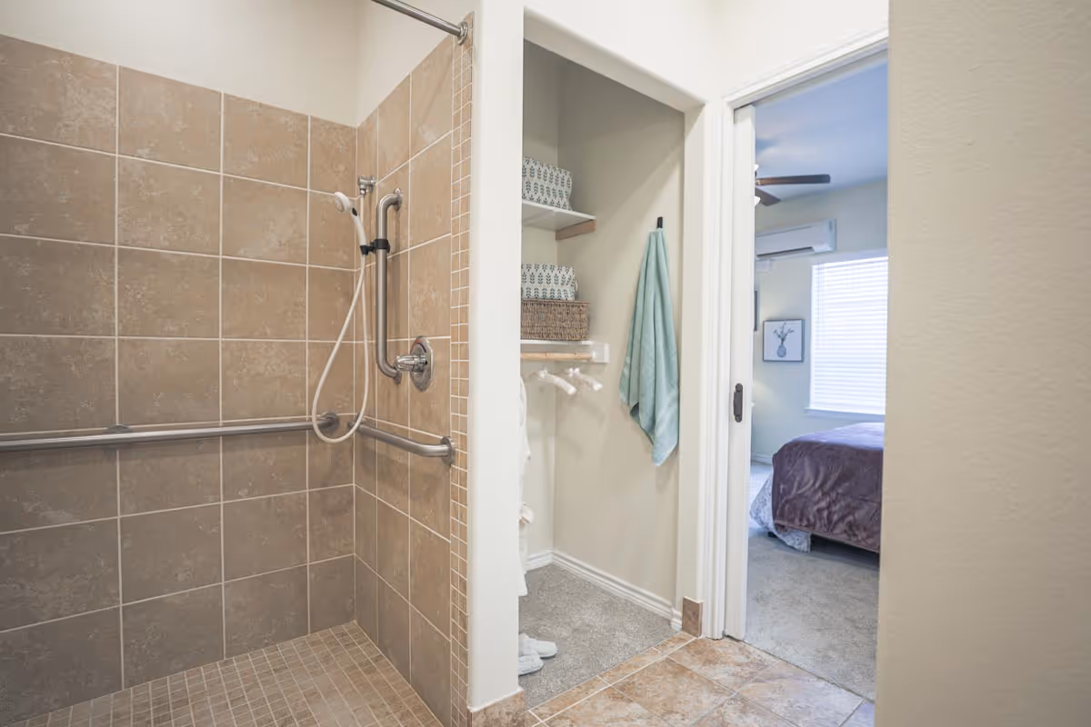 A tiled walk-in shower with grab bars and a handheld showerhead in a bathroom. Adjacent to the shower is a small closet with shelves holding towels and a hanging towel. Beyond the closet, there is a view into a bedroom with a bed, window with blinds, and a ceiling fan.