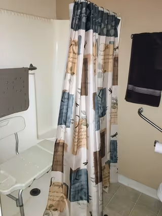 A bathroom shower area with a white shower chair inside the shower. The shower curtain has a decorative pattern with various colored blocks and cursive text. There are two towels hanging on the walls, one gray and one black. The floor is tiled and there is a toilet paper holder visible on the right side.
