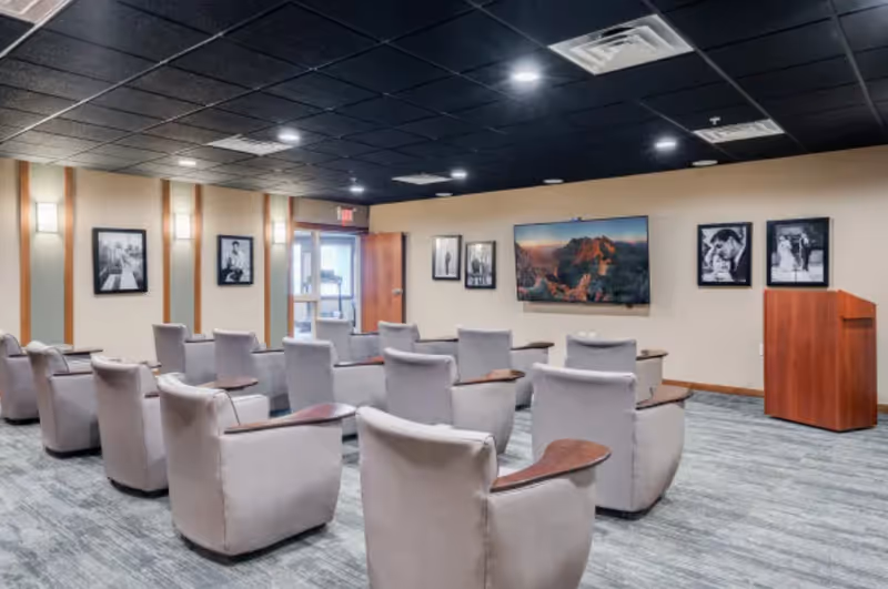 A small auditorium or presentation room with several rows of beige cushioned chairs with wooden armrests facing a wall-mounted flat screen TV displaying a mountain landscape. The room has a carpeted floor, beige walls adorned with black and white framed photographs, and a wooden podium on the right side. Ceiling lights illuminate the space.