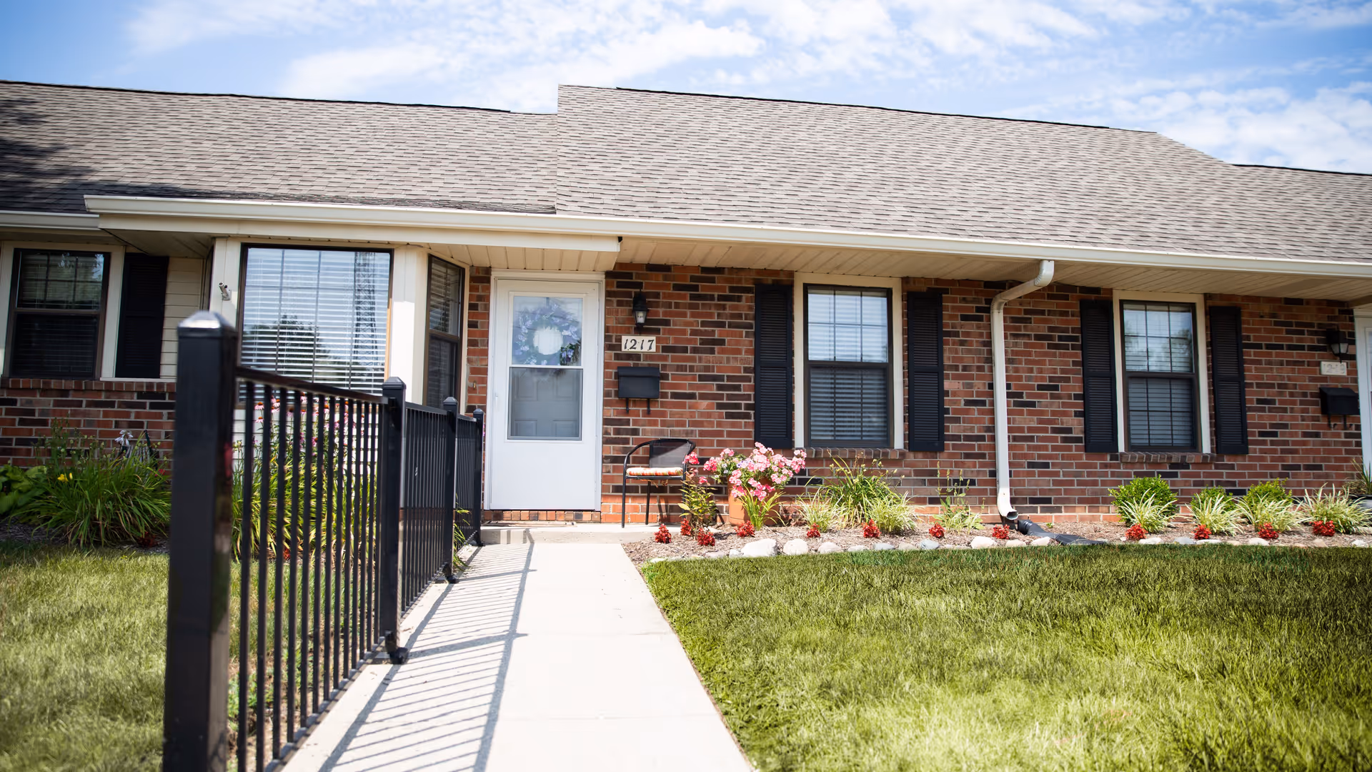 Exterior view of a single-story brick building with a white door and two windows with black shutters. A concrete walkway leads to the door, bordered by a black metal railing on one side and a landscaped area with green grass, plants, and flowers on the other side. The sky is partly cloudy.