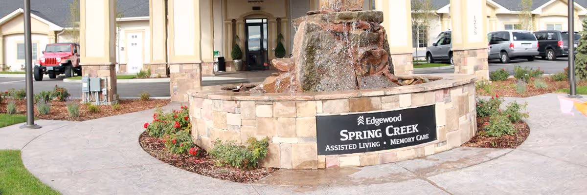 Outdoor view of the entrance area at Edgewood Spring Creek assisted living and memory care facility, featuring a stone fountain with water flowing and a sign displaying the facility name. Surrounding the fountain are landscaped flower beds and a paved walkway, with parked cars visible in the background.