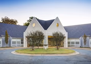 Front entrance of a nursing facility with a circular driveway, landscaped shrubs, and a peaked roof façade.