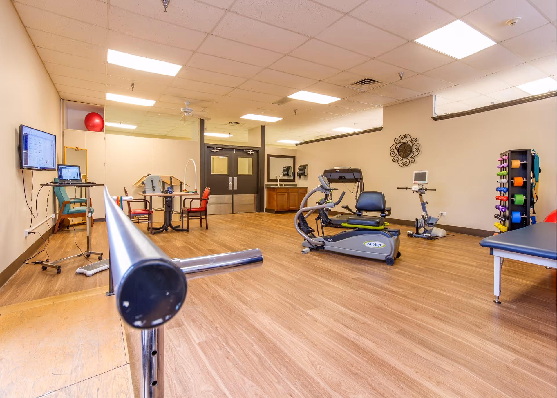 Bright rehabilitation/exercise room with parallel bars in the foreground, cardio machines, a weight rack and tables.