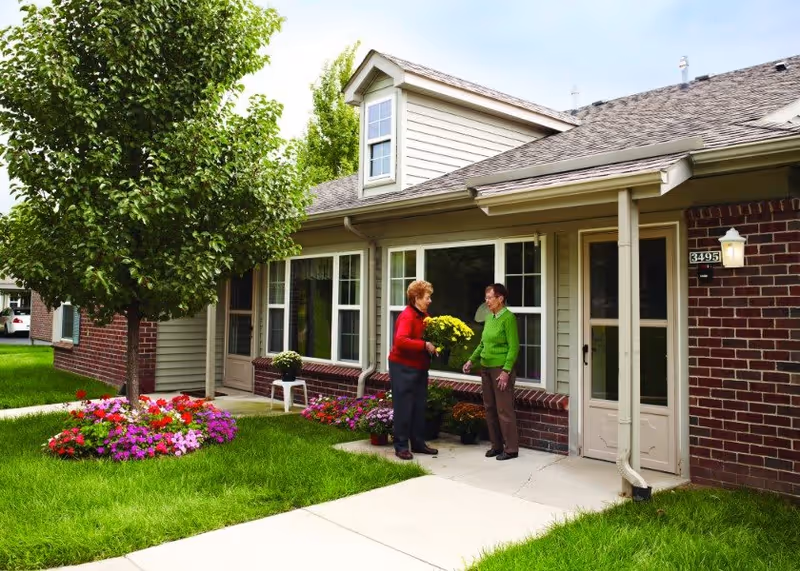 Two elderly women standing and talking outside a brick and siding building with large windows, surrounded by green grass, a tree, and colorful flower beds.