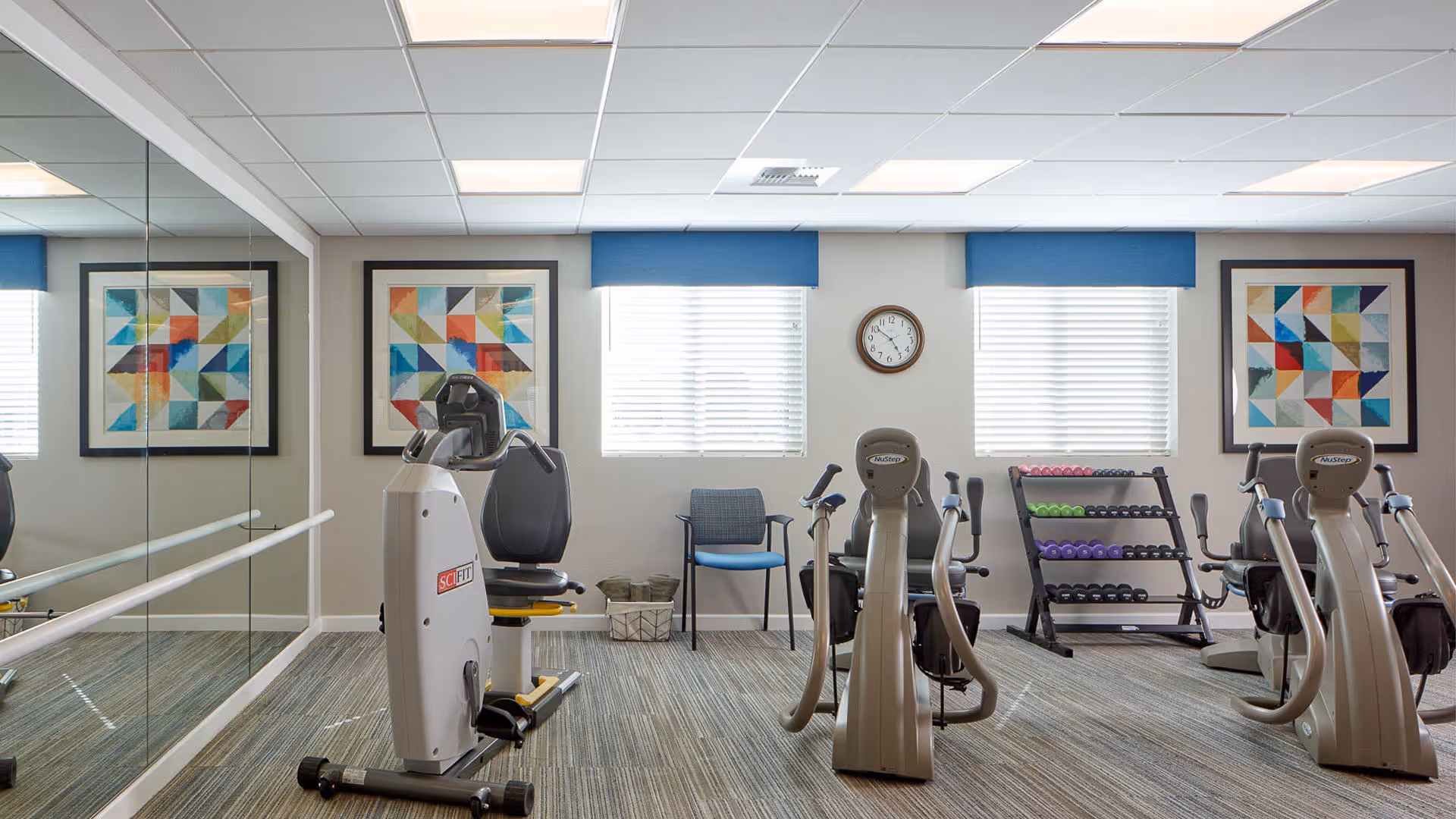A small fitness room with exercise equipment including recumbent bikes and a rack of colorful dumbbells. The room has two windows with blue valances, a wall clock, two framed abstract geometric art pieces, and a large mirror covering one wall.