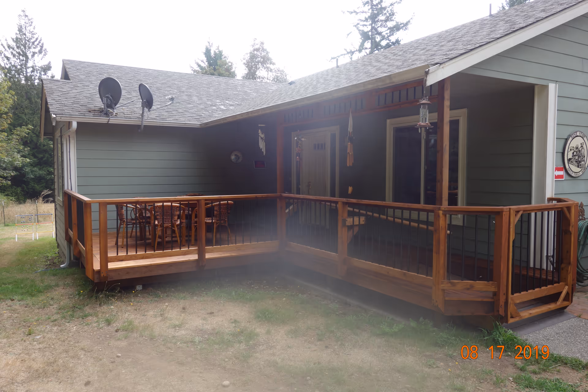 Wooden porch with railing and outdoor seating at the front of a single-story green house.