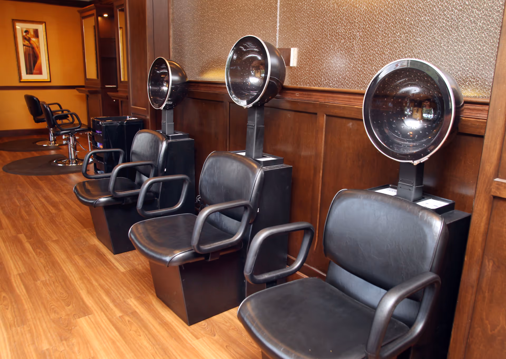 Interior view of a salon area with three black leather chairs each paired with a hair dryer hood. The room has wooden paneling on the walls, a wooden floor, and additional salon chairs and mirrors in the background.