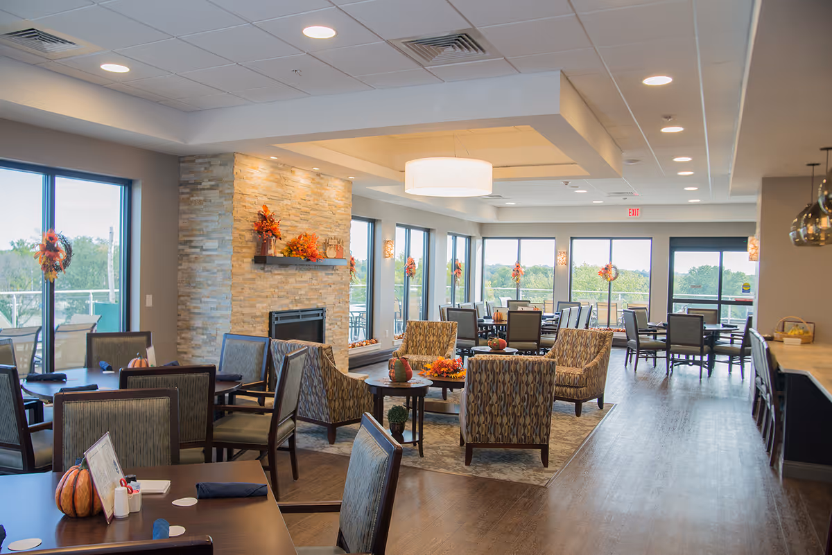 A spacious and well-lit common area in a senior living facility with multiple tables and chairs arranged for dining and socializing. The room features large windows with views of greenery outside, a stone fireplace decorated with autumn-themed floral arrangements, and comfortable patterned armchairs around a central coffee table. The ceiling has recessed lighting and a large hanging light fixture, and there is a counter with bar stools on the right side.