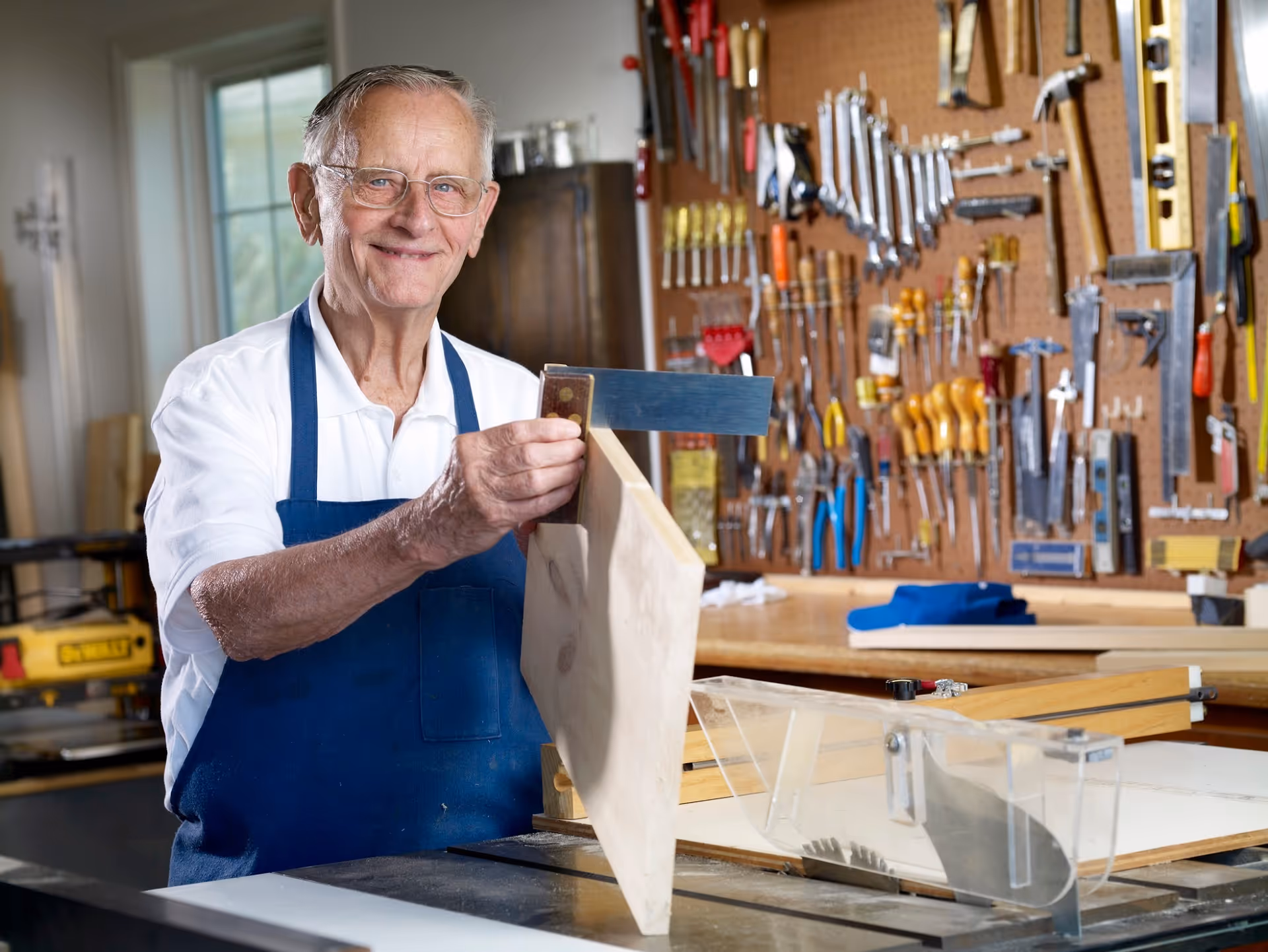 An elderly man wearing glasses and a blue apron is smiling while working with a wooden plank in a well-equipped workshop. Various tools are neatly organized on a pegboard behind him.