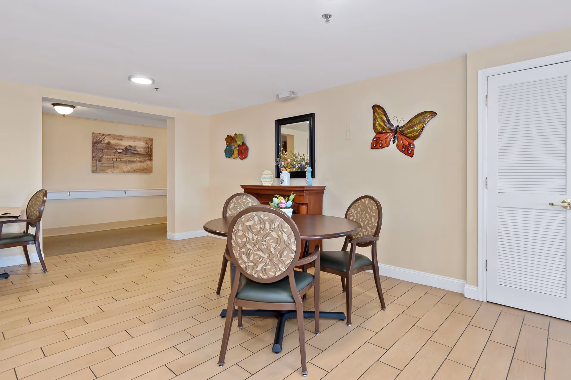 A small dining area in an assisted living facility with a round wooden table and four patterned chairs. The walls are beige with decorative items including a large butterfly wall art and a mirror above a wooden cabinet. The floor is tiled with light brown tiles, and there is a white door on the right side. In the background, there is a hallway with a framed picture on the wall.