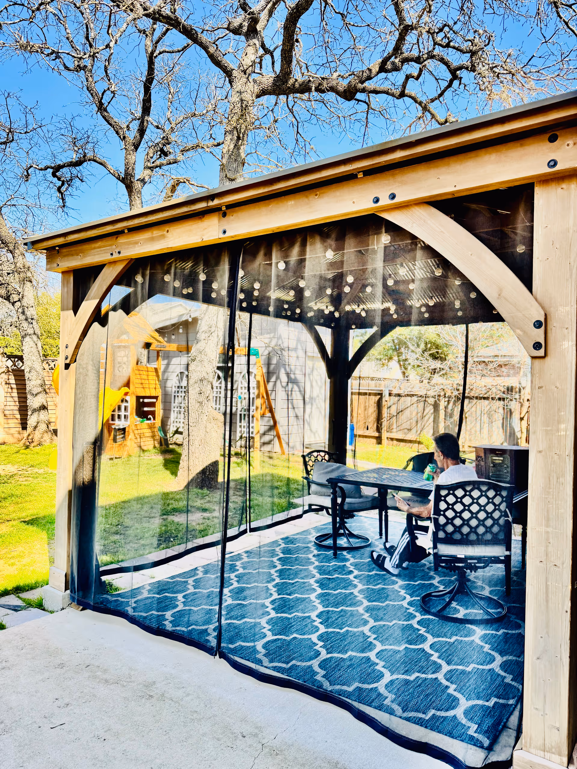 A person sitting on a chair at a table inside a wooden gazebo with clear plastic curtains. The gazebo is set on a blue patterned outdoor rug and is located in a backyard with trees, a wooden fence, and a play structure visible in the background.