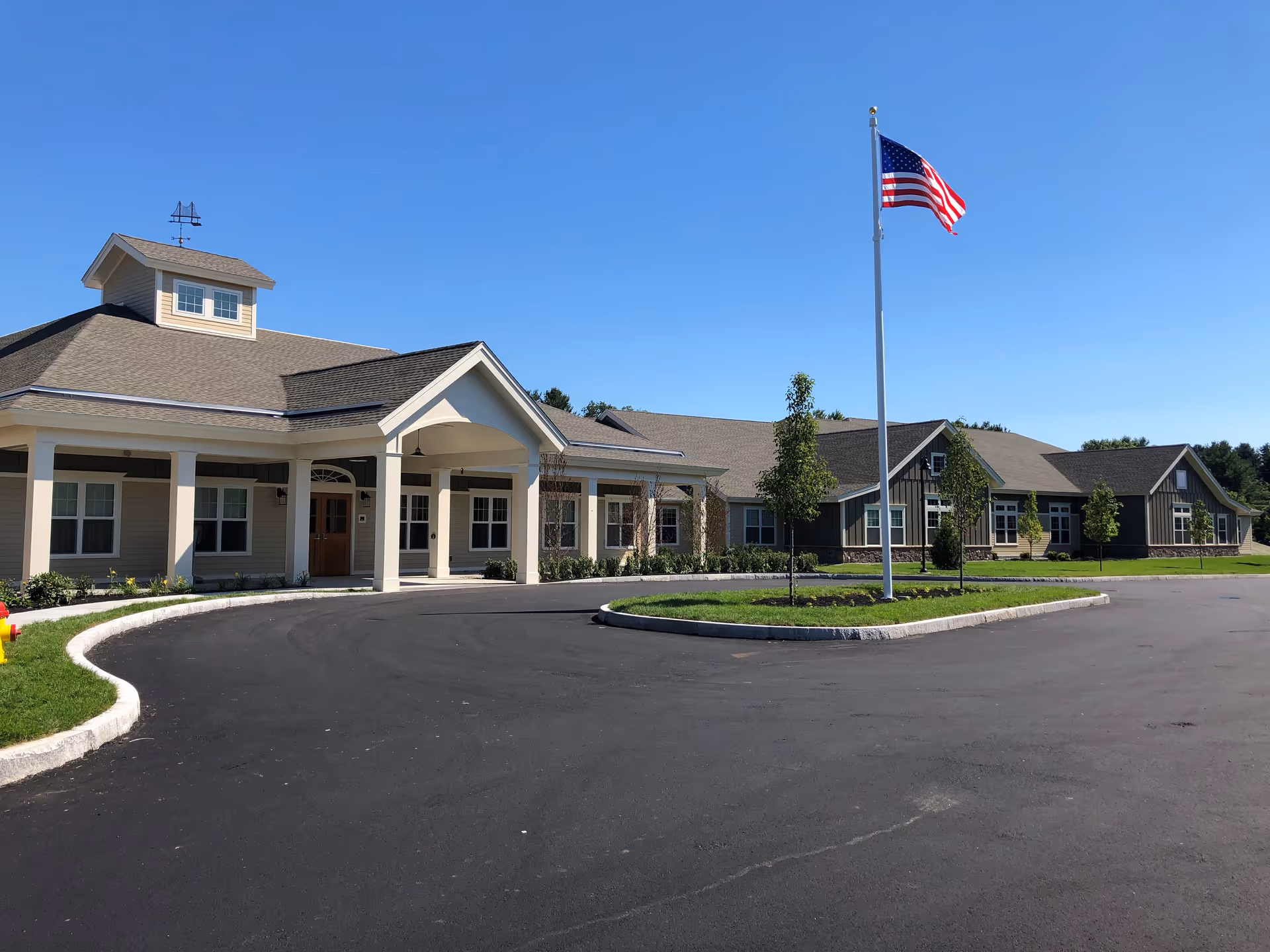 Single-story senior living facility building with a covered entrance, circular driveway, and an American flag on a flagpole.