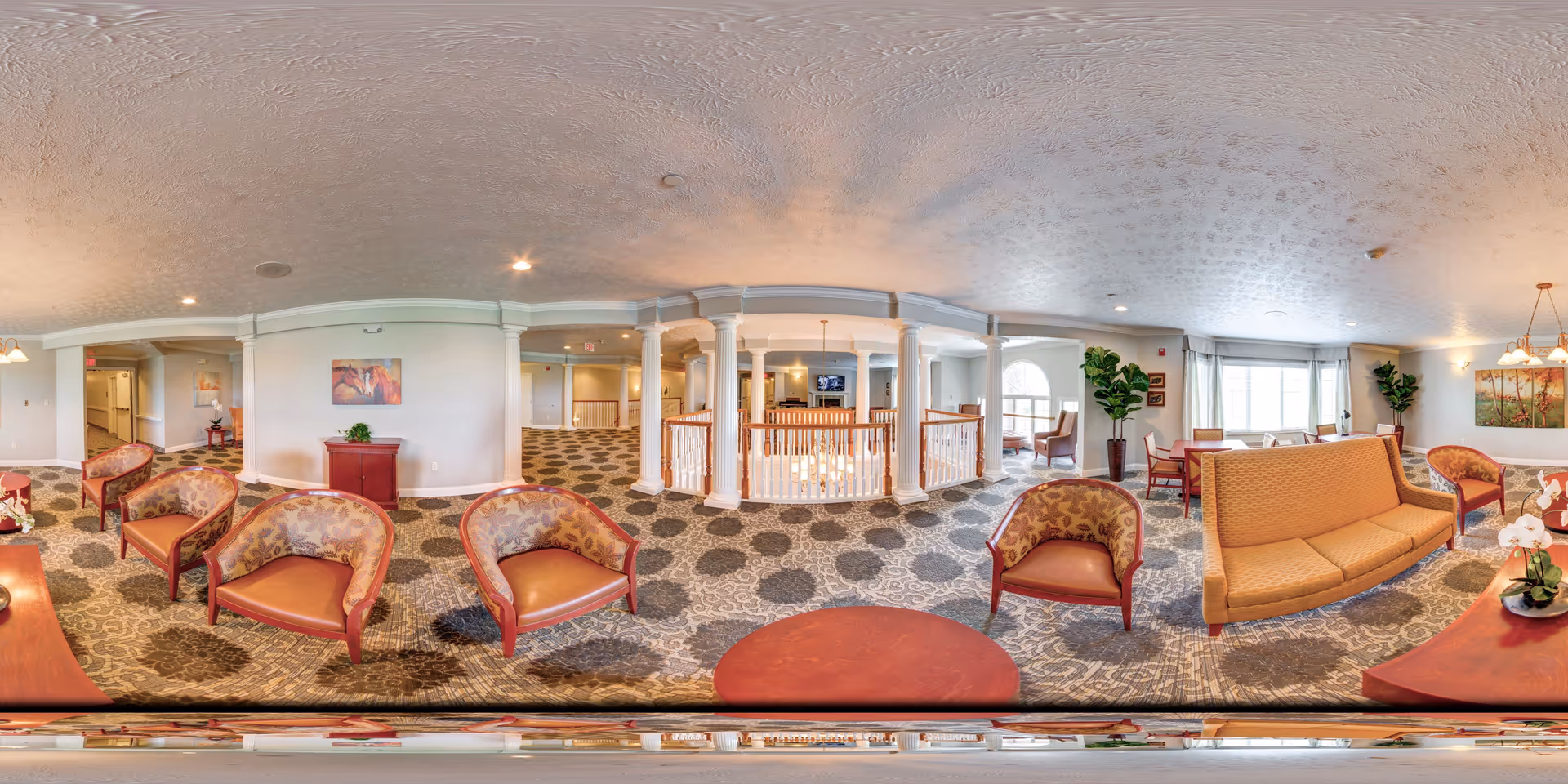 A spacious and well-lit common area in a senior living facility featuring patterned carpet flooring, multiple upholstered chairs and sofas arranged around wooden tables, decorative plants, and wall art. The area includes white columns and railings overlooking a lower level, with large windows allowing natural light to fill the room.