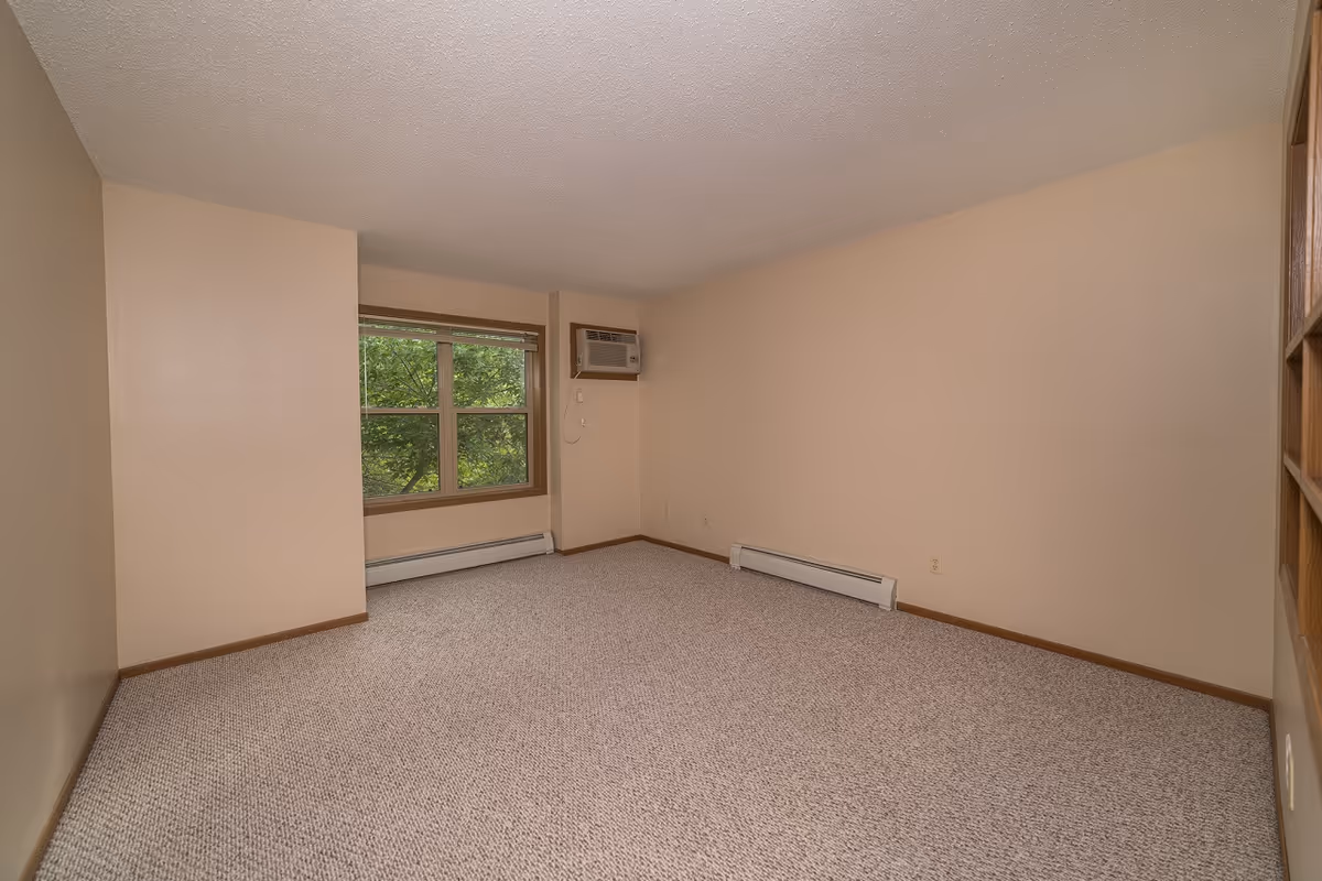 Empty room with beige walls and carpeted floor, a window with a view of green trees, and a wall-mounted air conditioning unit.