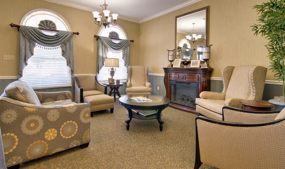 Cozy common living room with upholstered chairs and sofa arranged around a round coffee table and a fireplace beneath a large mirror.