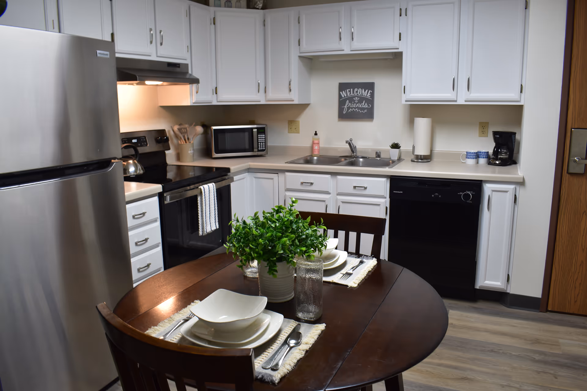 A modern kitchen with white cabinets, stainless steel refrigerator, black stove, microwave, dishwasher, and a coffee maker. A round wooden dining table is set with white dishes, glasses, silverware, and a green potted plant centerpiece. A small sign on the wall above the sink reads 'Welcome friends'.